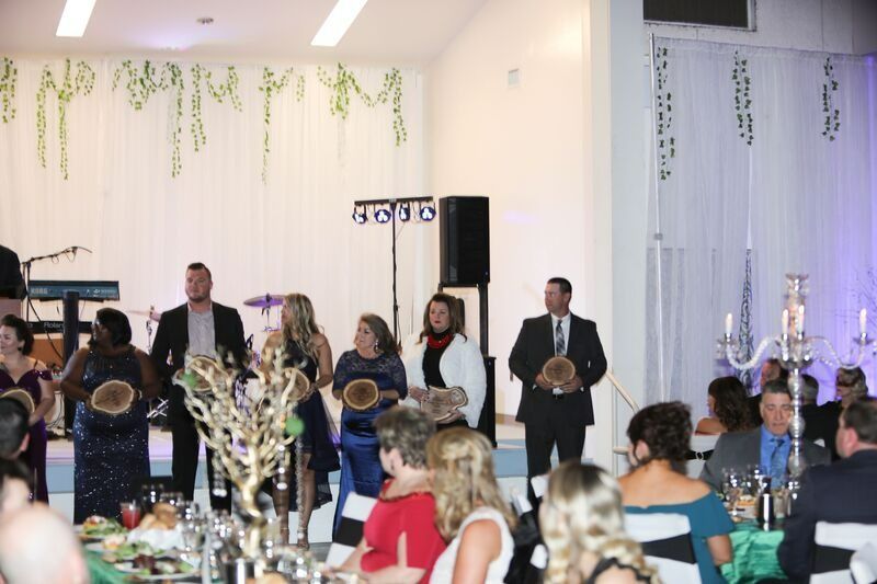 People at an awards ceremony, holding plaques.  Stage backdrop with greenery and lights.  Audience seated at tables.