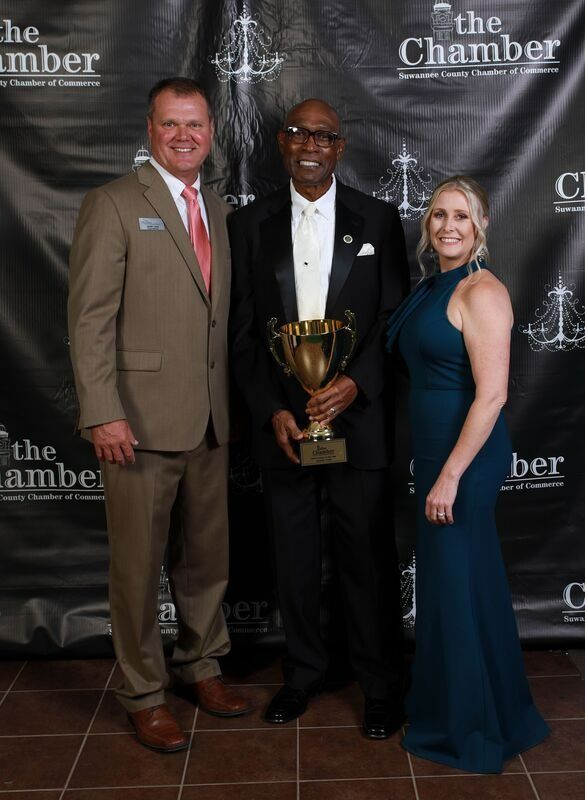 Three people posing with an award at a formal event. Man in tuxedo holds trophy, flanked by others.