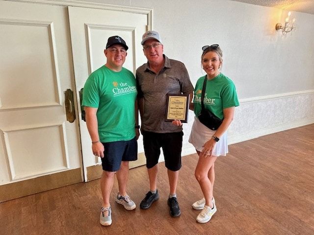 Three people pose indoors: two in green shirts with a man holding an award.