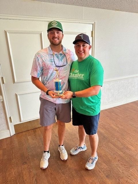 Two men holding a trophy; one in a floral shirt, the other in a green shirt. Indoors, smiles.