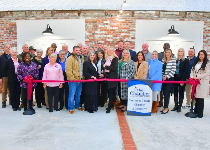 Group cuts ribbon at an event hosted by The Chamber. Brick building, pink ribbon, many people smiling.