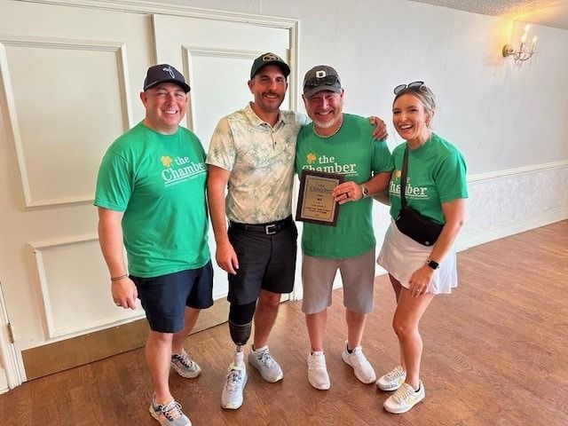 Four people in green shirts pose with a plaque indoors. One man has a prosthetic leg.
