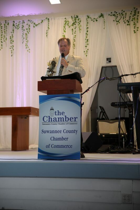 Man speaking at Chamber of Commerce podium. White suit, microphone, floral background, event setting.