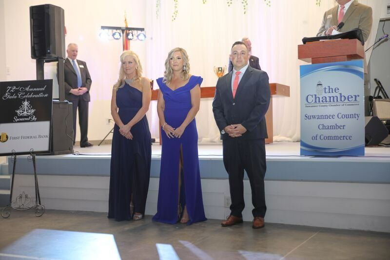Three people stand on a stage with a podium and Chamber of Commerce sign.