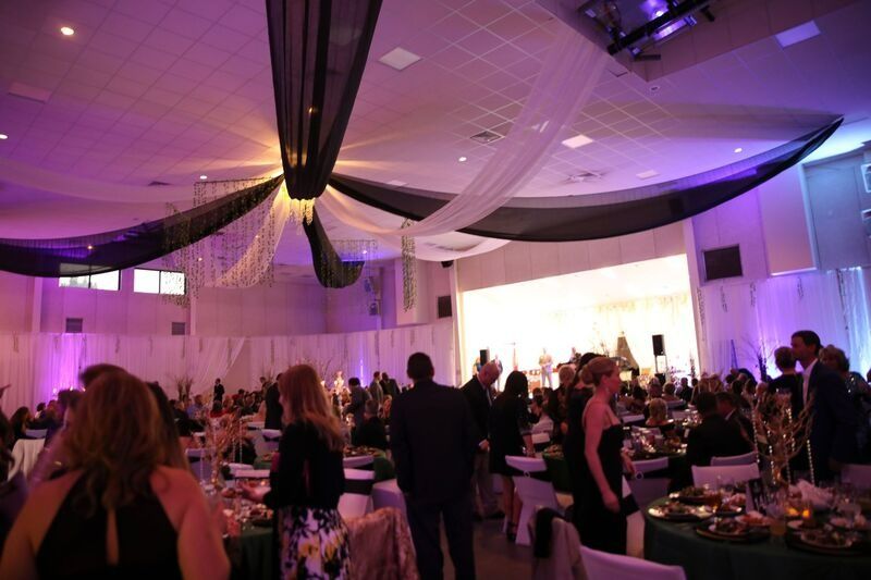 Event in a decorated hall with people mingling. Black and white draped ceiling, tables set, purple lighting.
