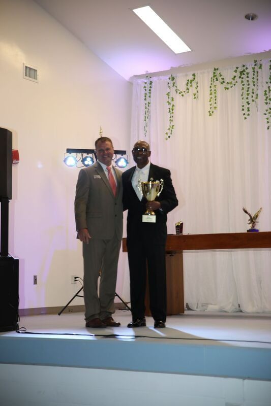 Two men on a stage; one presents an award to the other. Formal attire, neutral expressions, indoor setting.