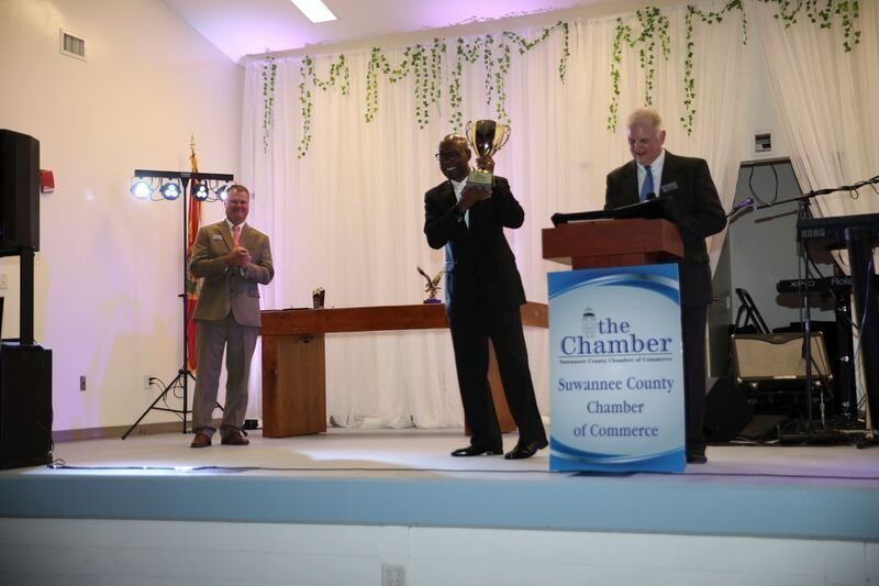 Man in suit holding trophy on stage, flanked by two men, at an awards ceremony.