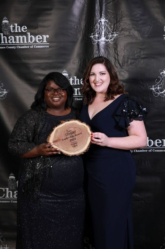 Two women holding award, smiling in front of a backdrop with logos. The award is a wood slice.