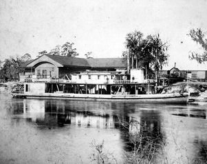 Paddle steamer boat on a river, with a large building and trees in the background.