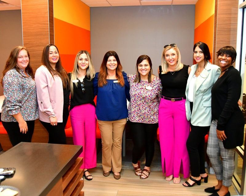 Group of eight people posing together indoors, some in pink pants.