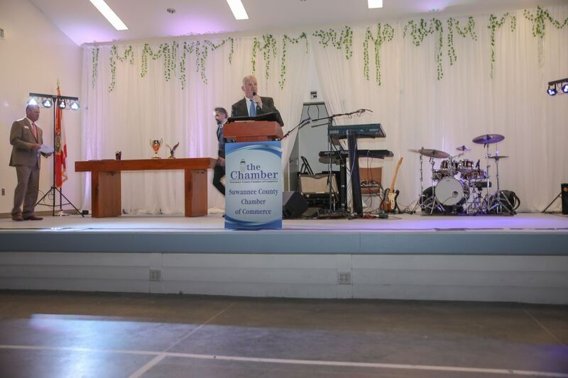 Man speaking at podium on stage with band instruments and backdrop.