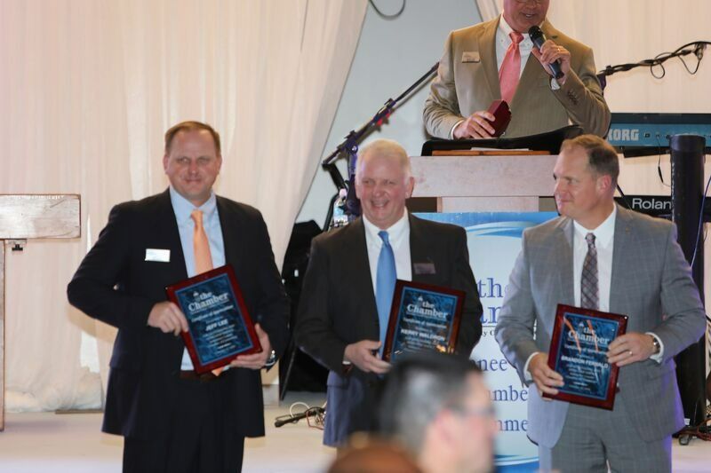 Three men in suits holding awards, standing at a podium. A man speaks into a microphone behind them.