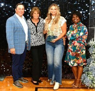 Four people stand together; a man in blue blazer, two women, and another woman in floral dress.