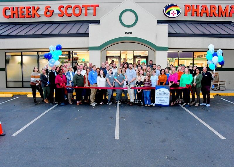 People at Cheek & Scott Pharmacy ribbon-cutting. Many people standing, holding ribbon in front of building. Balloons present.