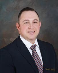 Man in suit smiling, looking at camera. Navy jacket, patterned tie, white shirt, against a dark background.