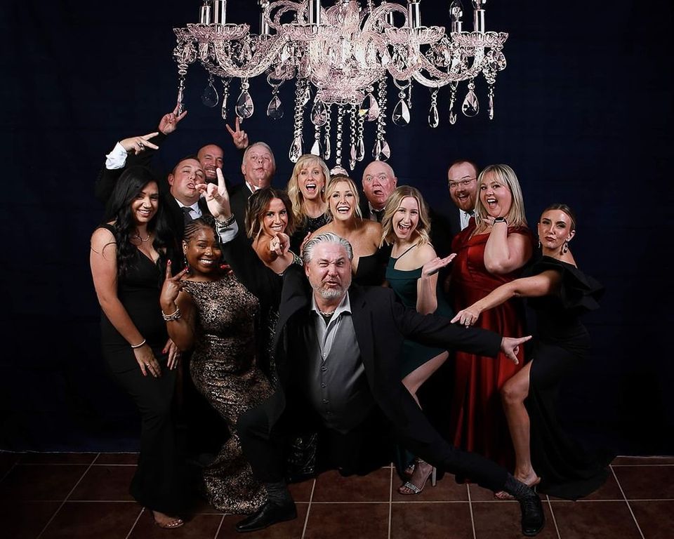 Group of people in formal attire pose under a chandelier, some making playful gestures. Dark background.