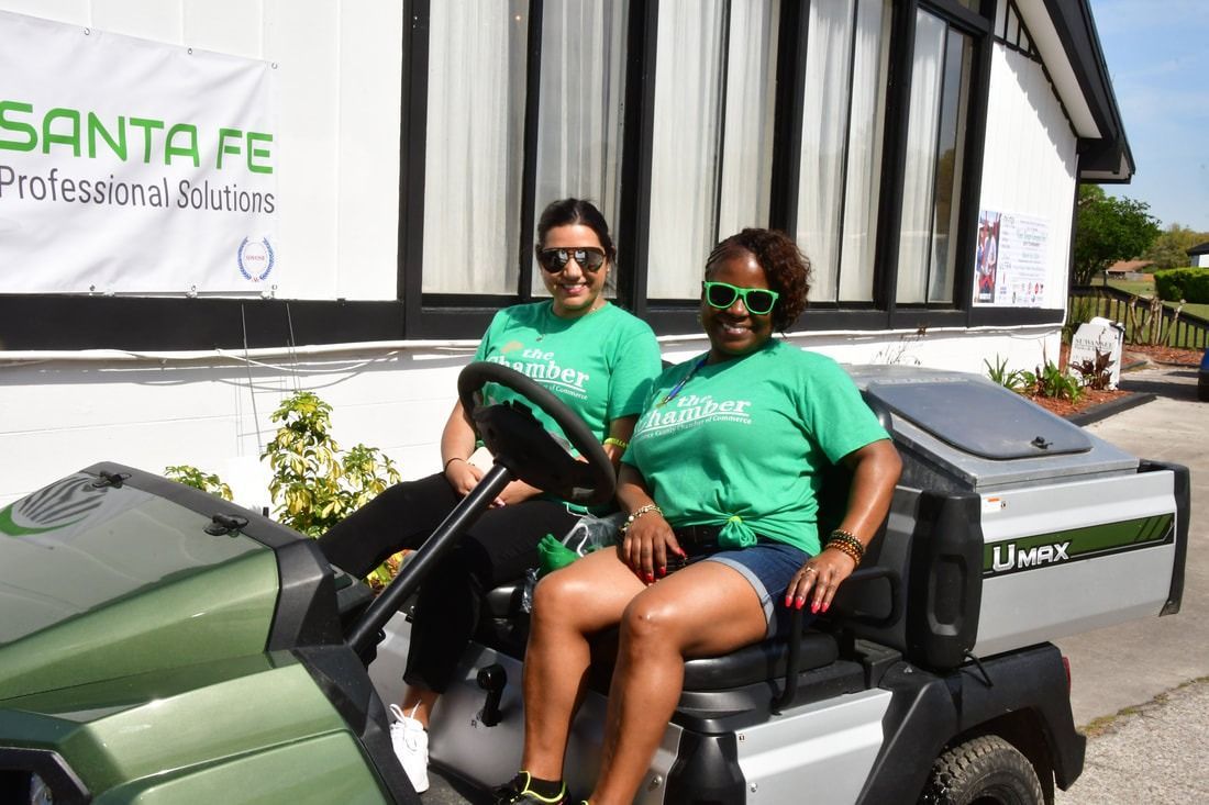 Two women in green shirts sit in a green golf cart. They smile. Building in the background.