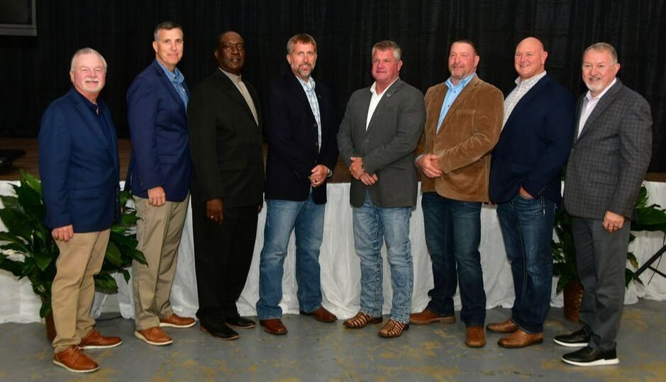 Group of men in business attire, standing indoors in front of a backdrop, smiling.