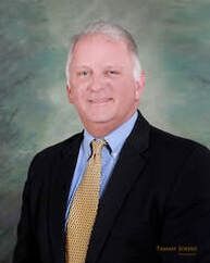 Man in suit, smiling, with light blue shirt and yellow tie, in front of a green and blue background.