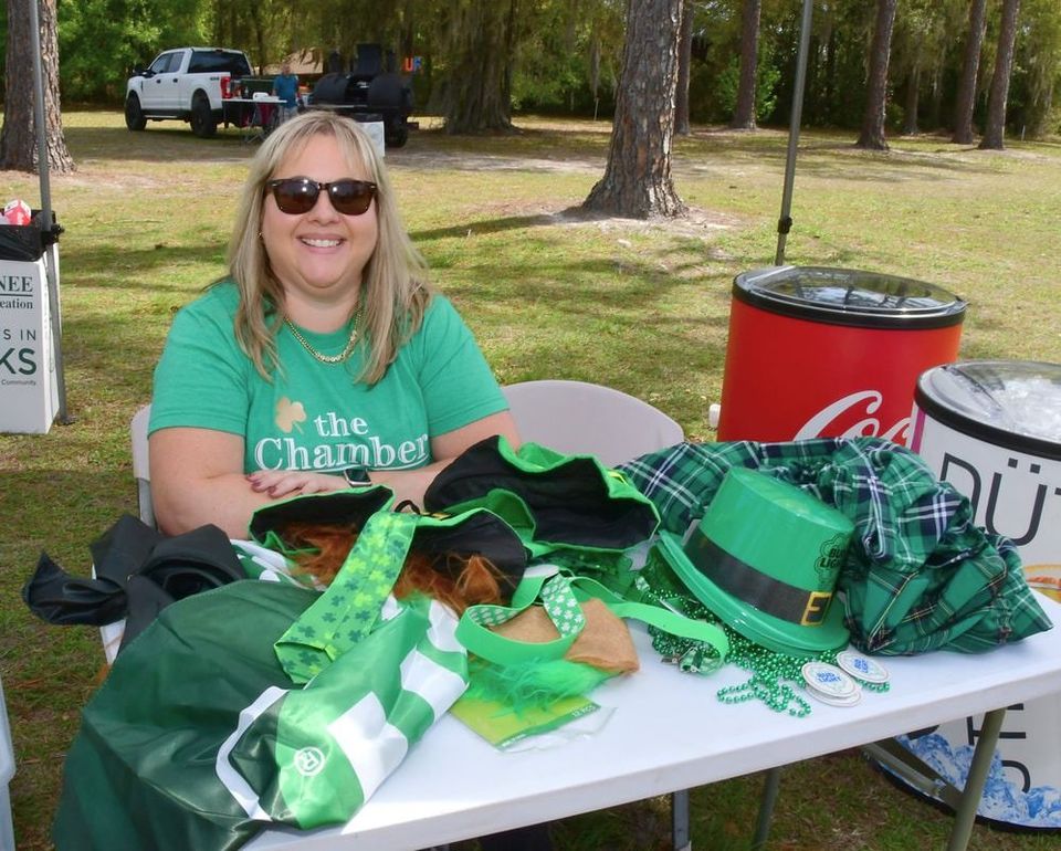 Woman in green shirt and sunglasses at a table with St. Patrick's Day merchandise outdoors.