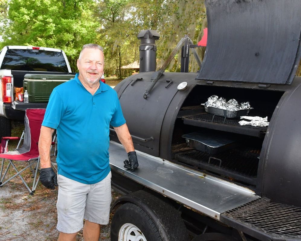 Man standing next to a large black smoker on a trailer, wearing gloves.
