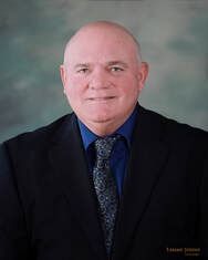 Man in a suit smiles at the camera, with a blue shirt and patterned tie.