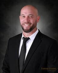 Man in black suit smiles at the camera, dark background.