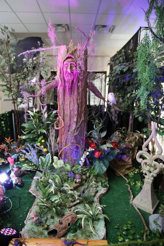 A whimsical forest display featuring a carved tree with a face, surrounded by plants and fairy lights.