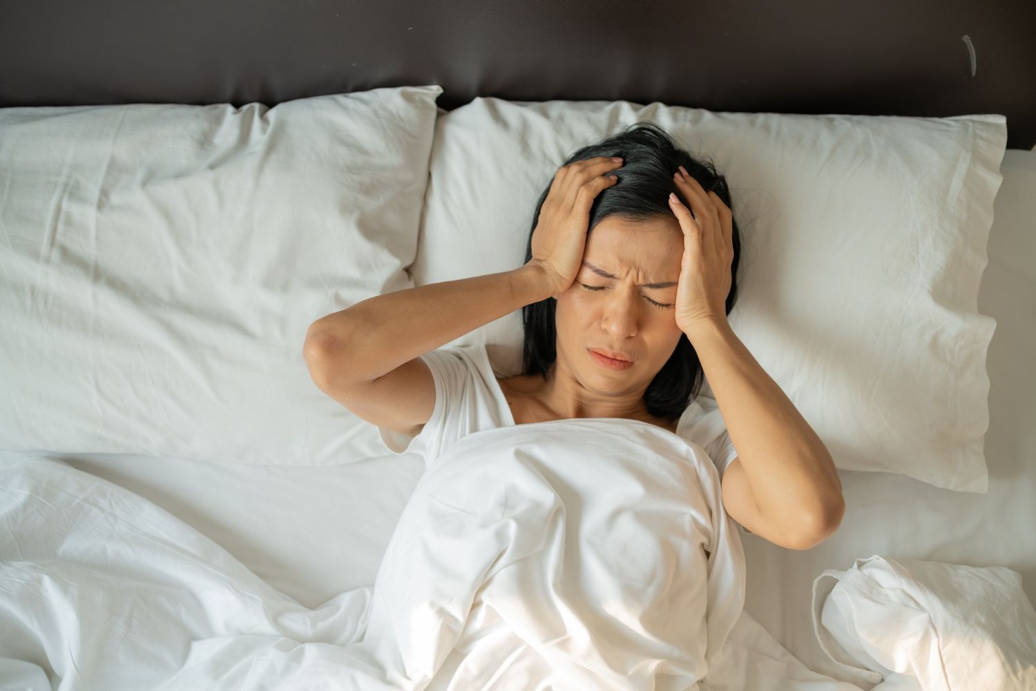 Woman in bed with hands on head, eyes closed, indicating headache.