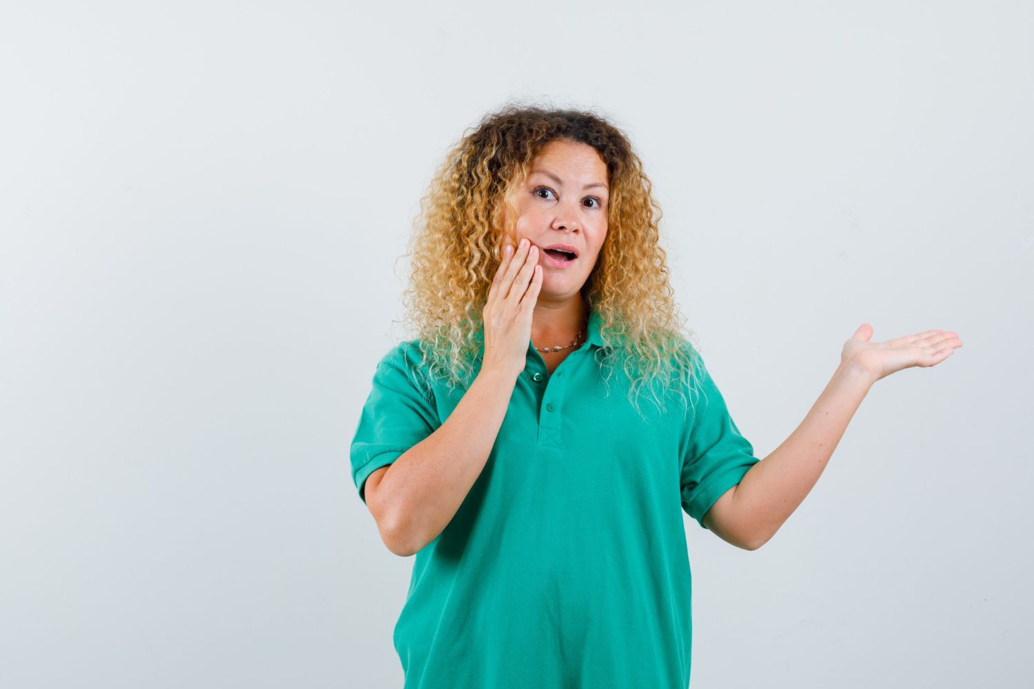 Woman with curly hair, wearing a green polo shirt, looks surprised, hand on cheek, arm extended.
