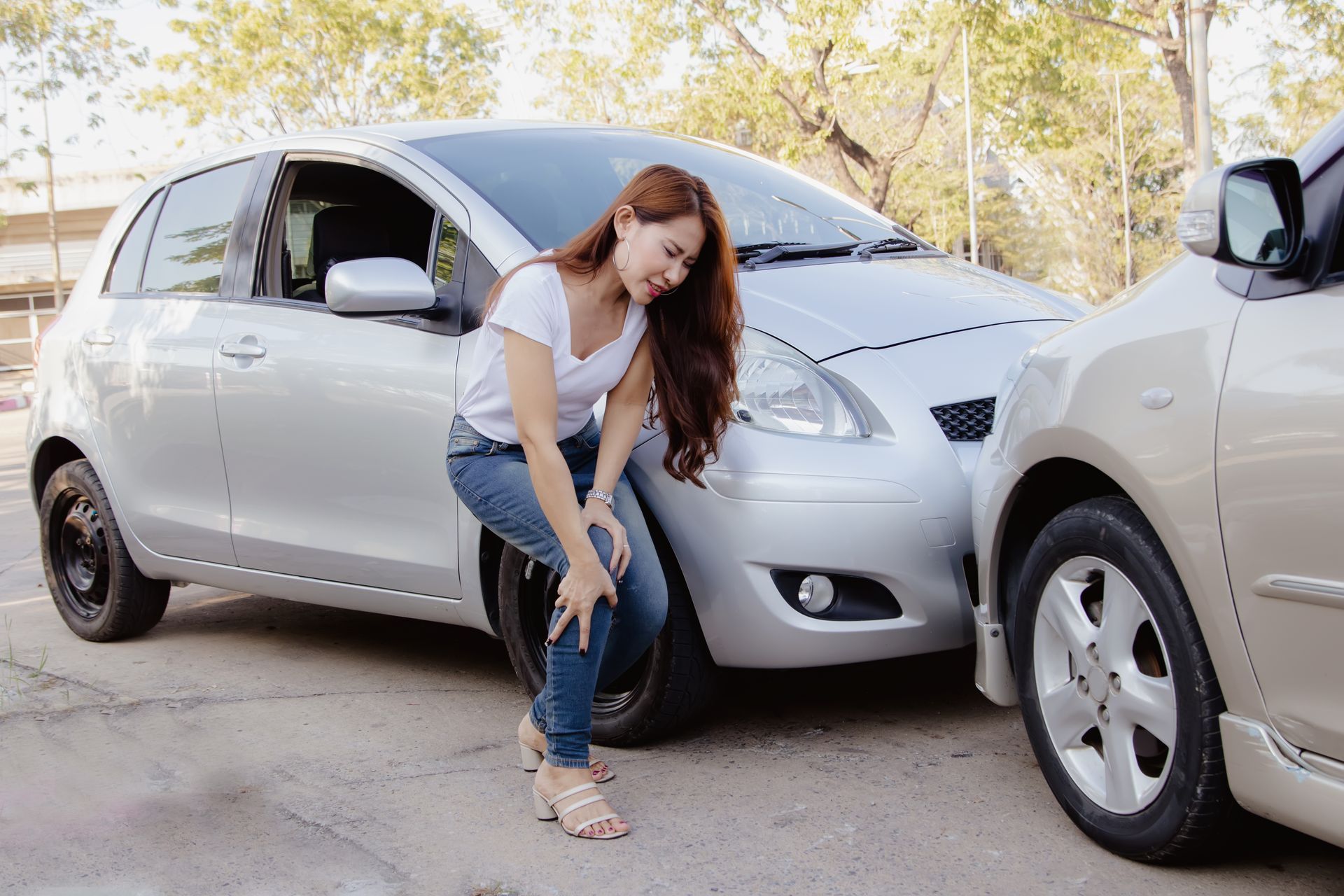 Woman crouching beside a silver car in a parking area, with another car nearby.