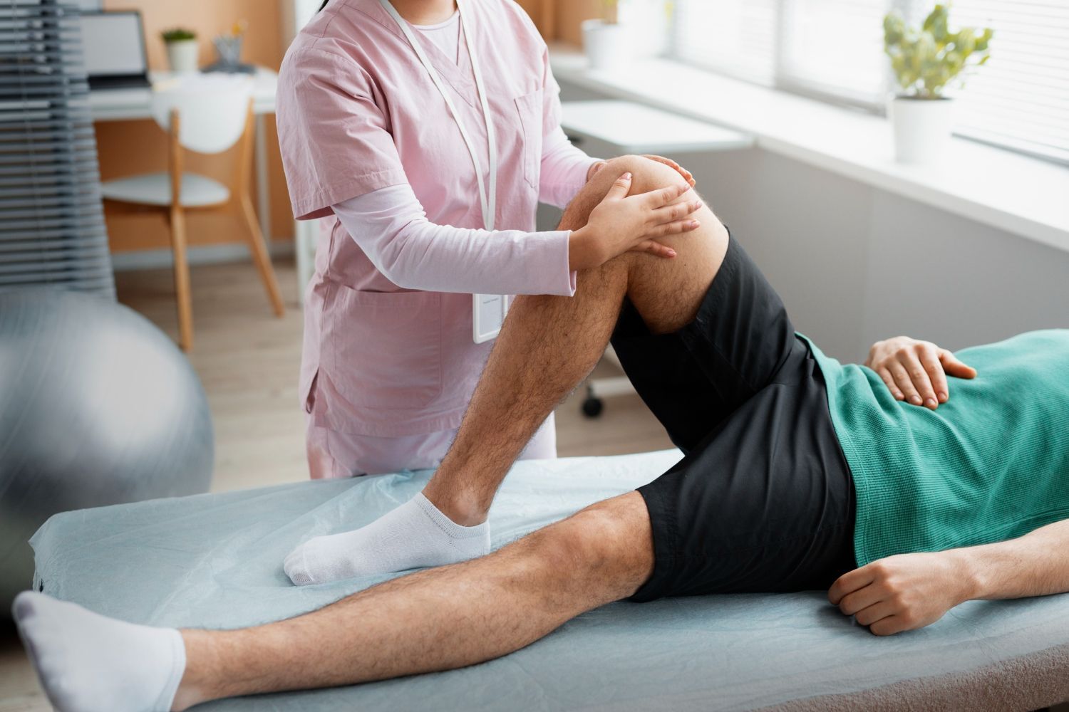 Physiotherapist examining a patient's knee in a bright room. Patient lies on a table, wearing shorts and a green shirt.