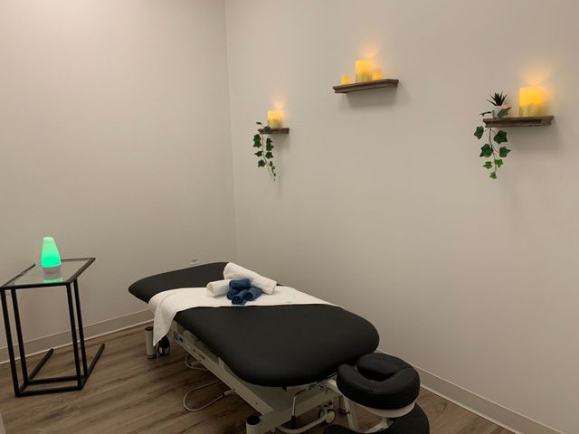 Spa treatment room with black massage table, folded towels, candles, and shelves on a beige wall.