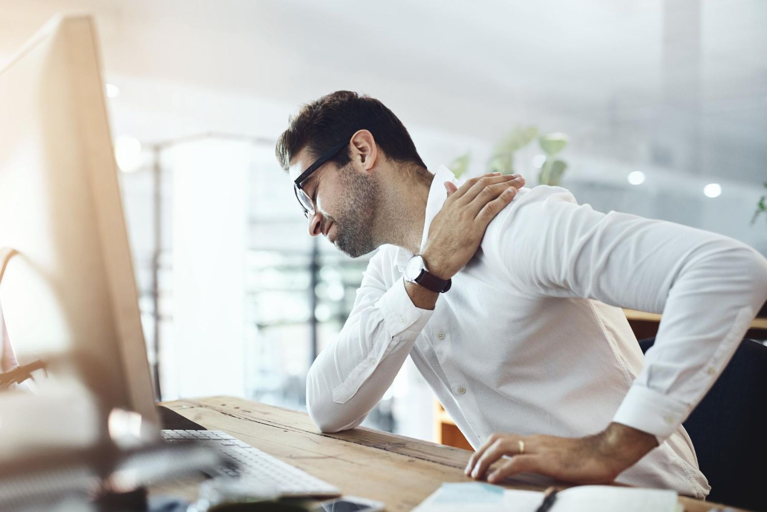 Man at desk with shoulder pain, touching his shoulder, at a computer.