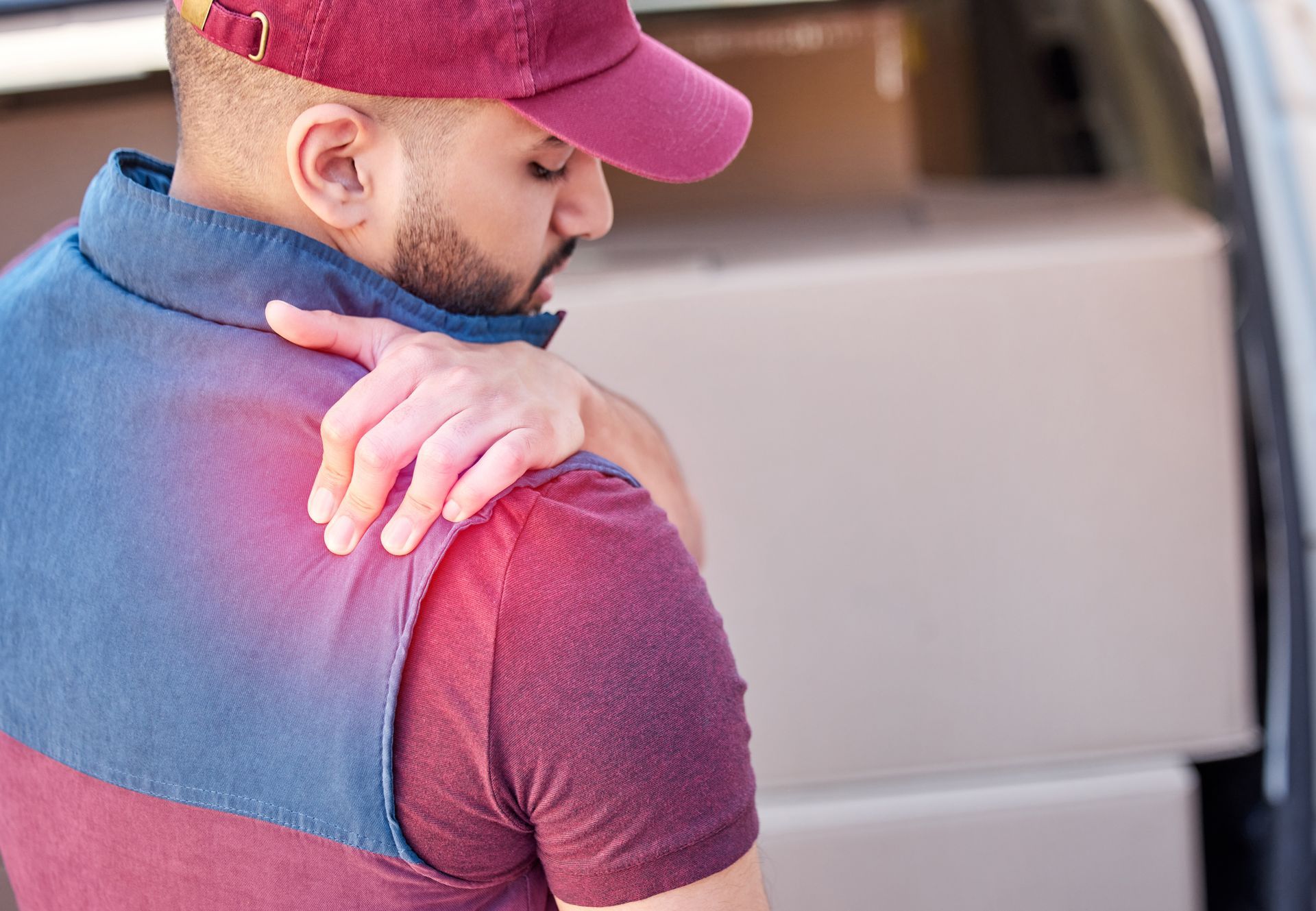 Man in red cap holding his shoulder, with a bright light on his upper back near a car trunk