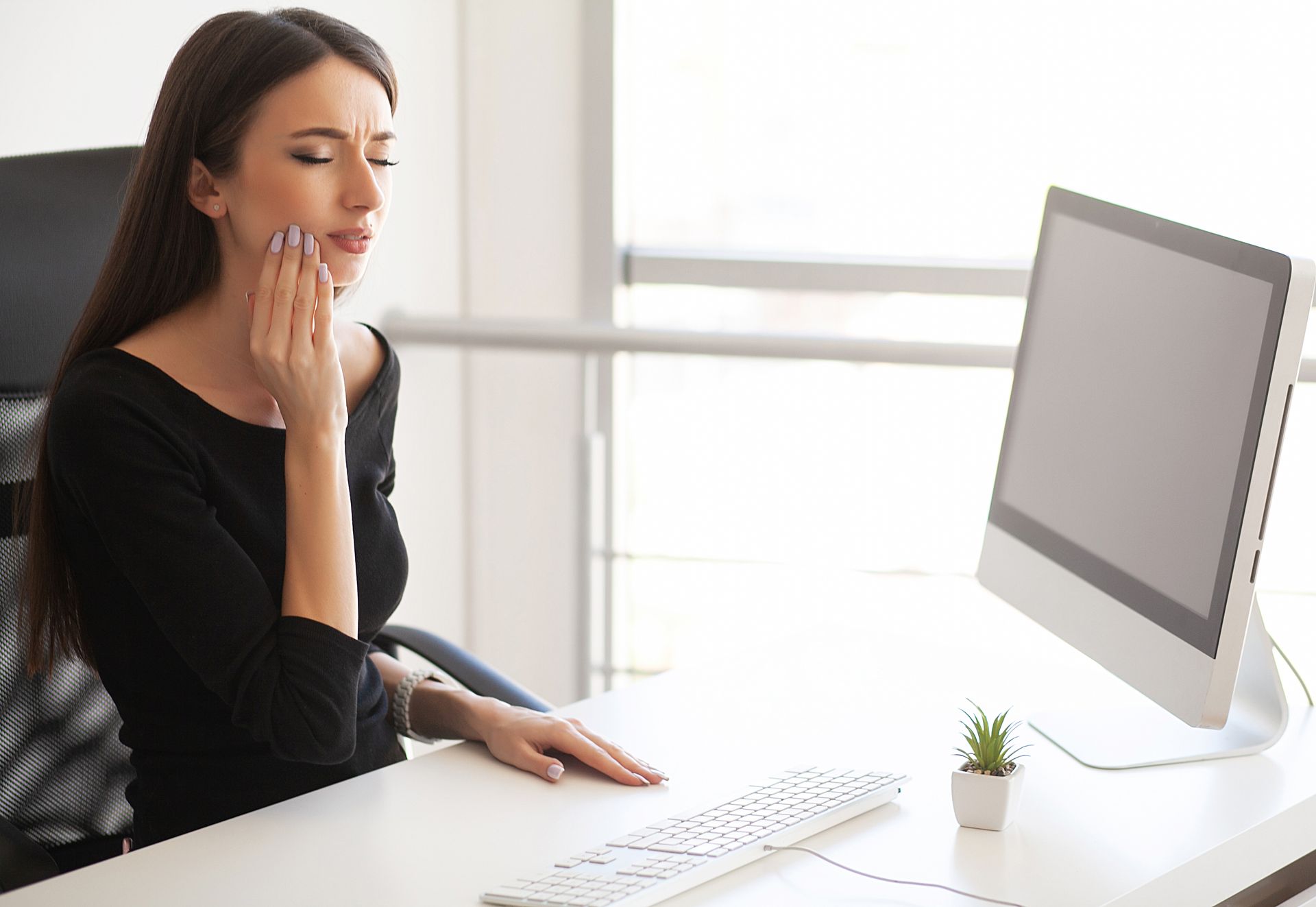 A woman is sitting at a desk with a computer and has a toothache.