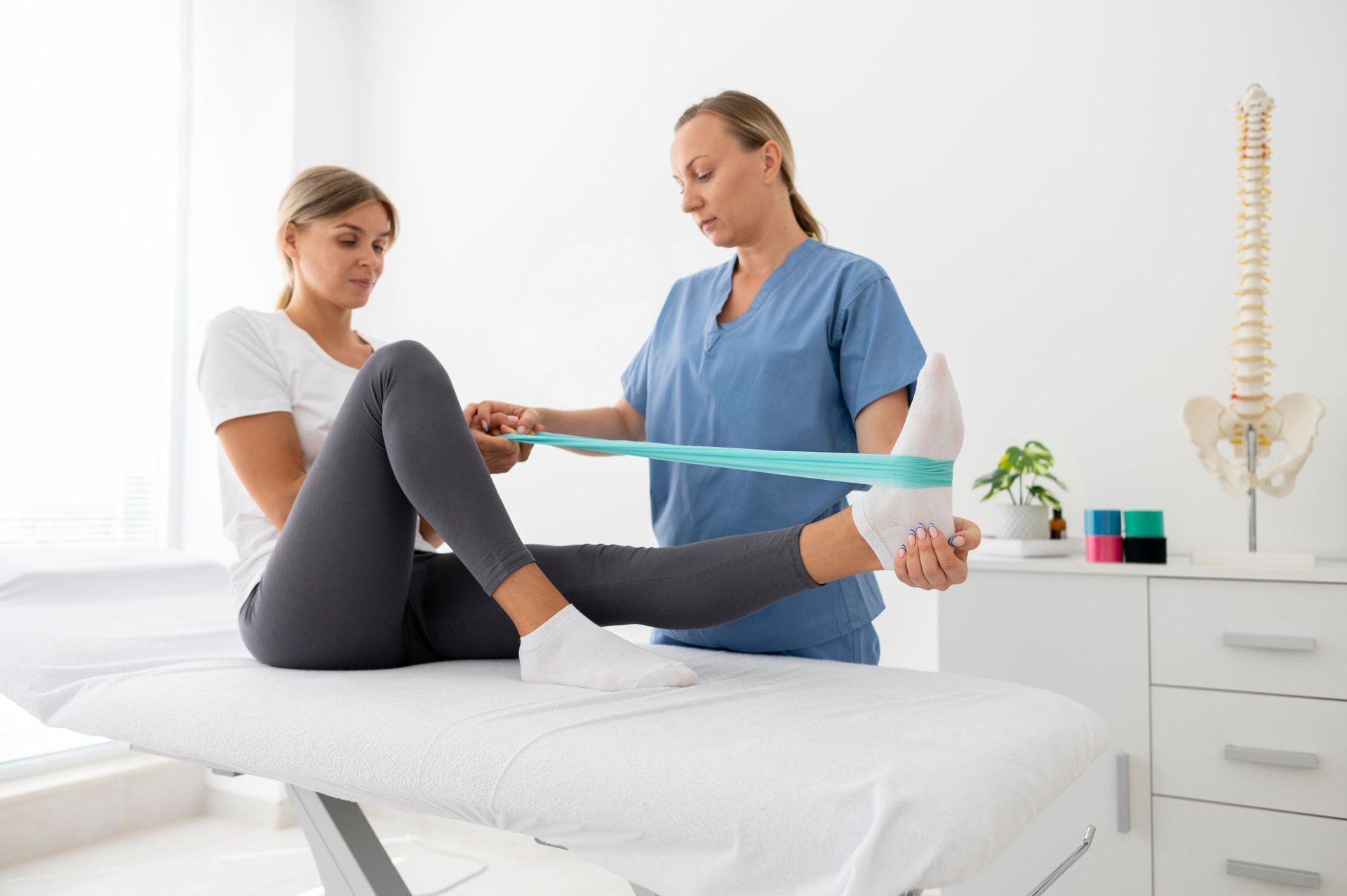 A woman is sitting on a bed while a nurse stretches her leg with a resistance band.