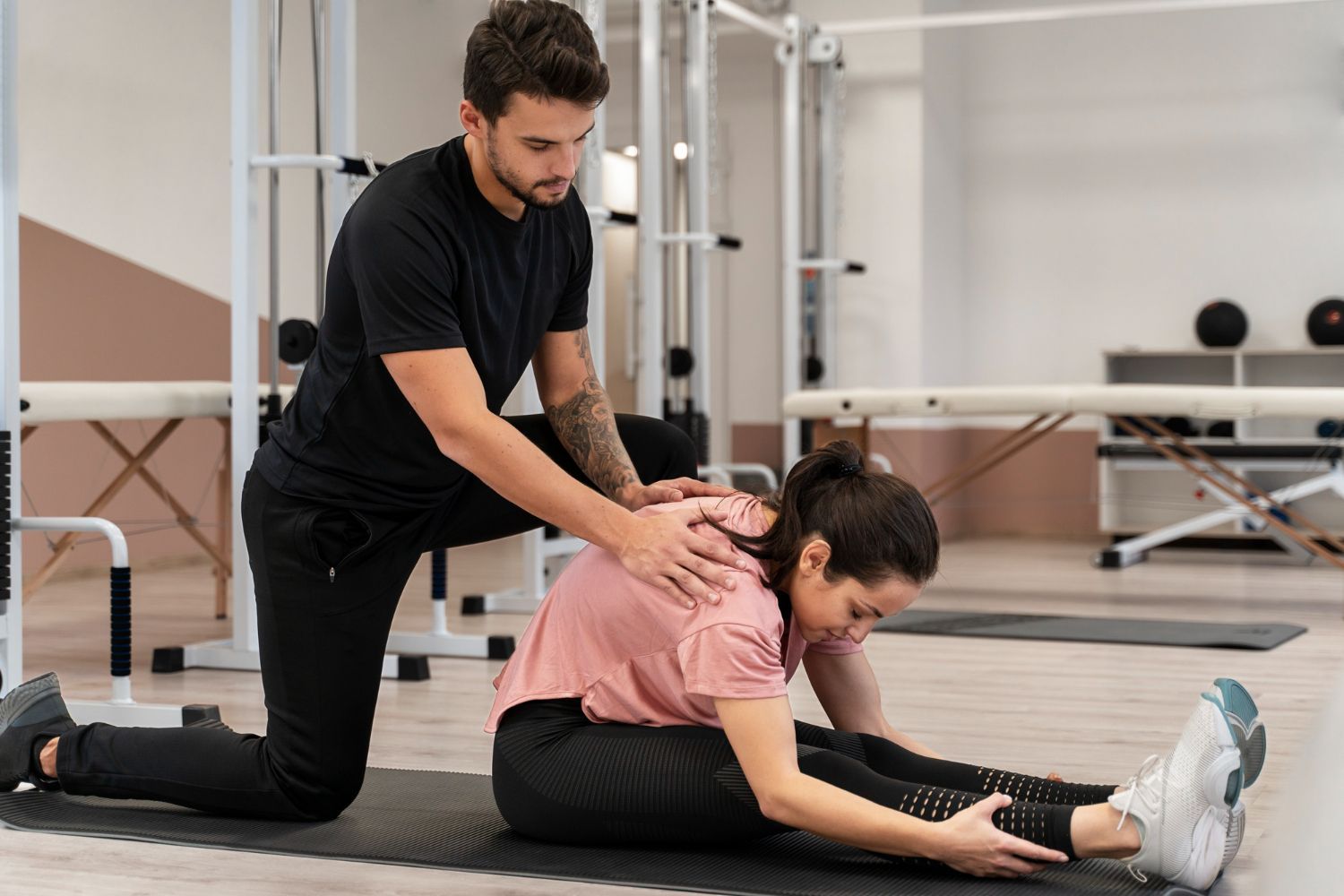 A man is helping a woman stretch her legs in a gym.