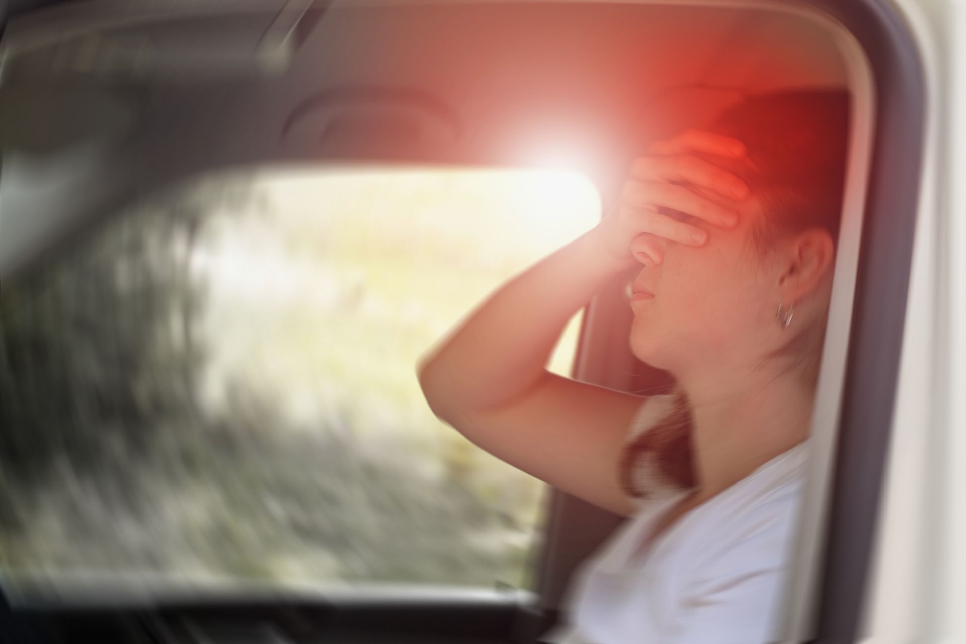 Woman in car, holding head with hand, red highlight indicating pain.