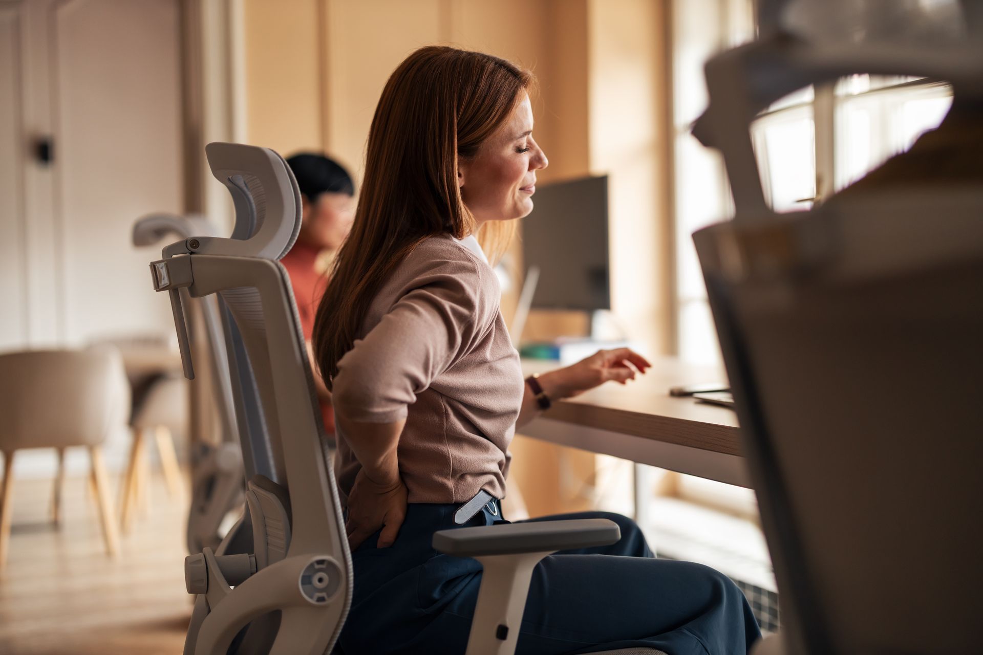 Woman in office chair, clutching her lower back, looking pained. Another person and desk visible in background.