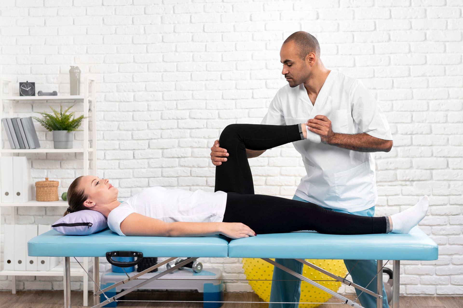 A physical therapist assisting a woman with a leg stretch on an examination table.