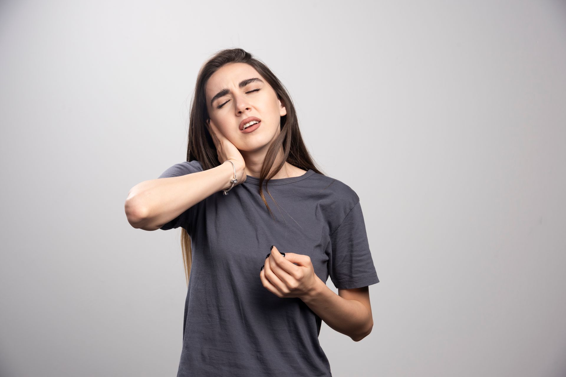 Woman holding her jaw in pain, gray shirt, light background.