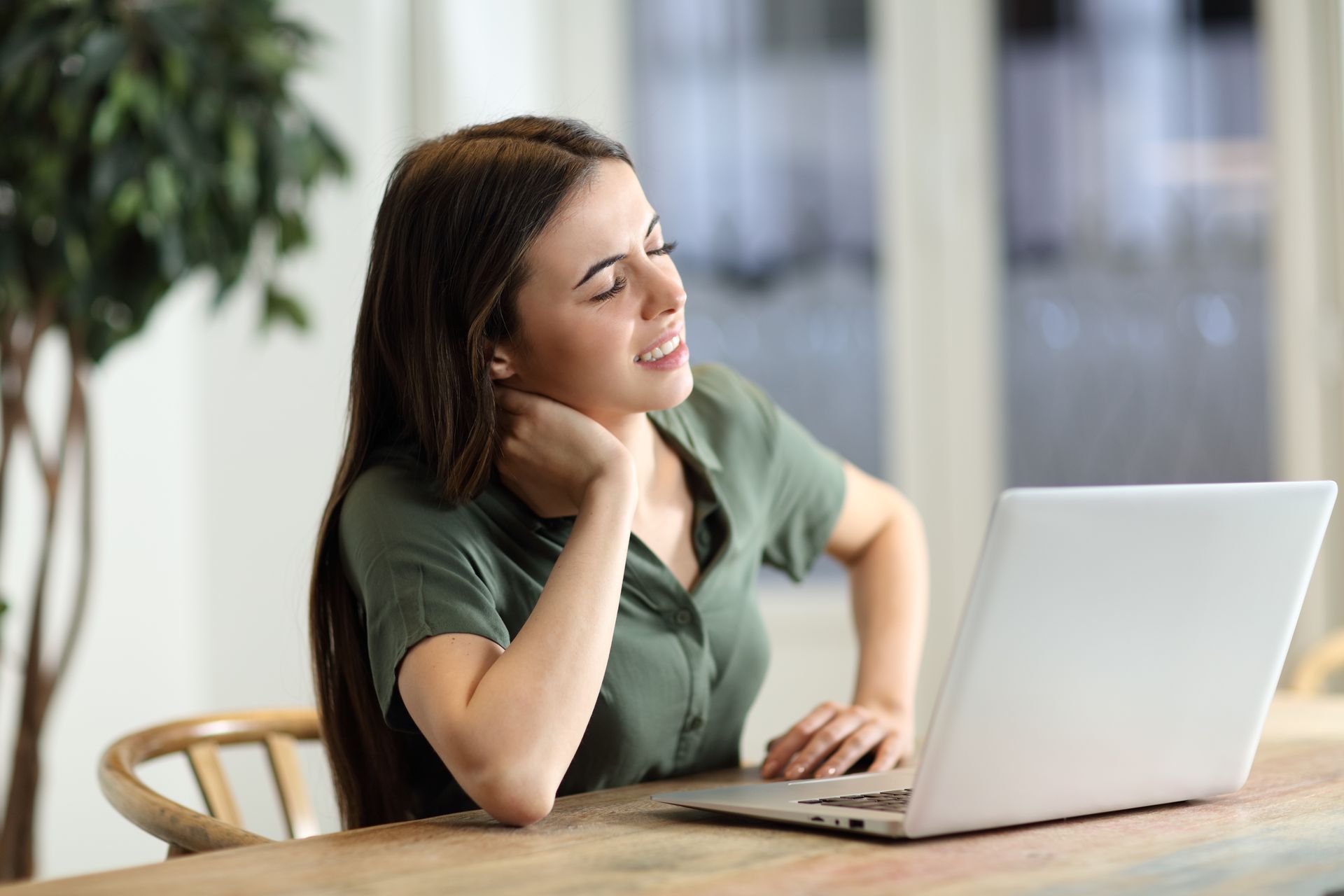 Woman holding her neck while working on a laptop at a table, looking pained.