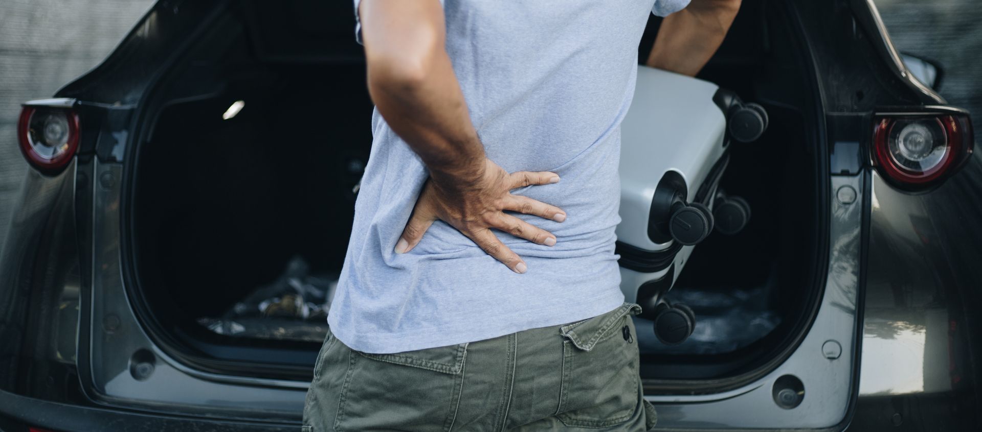 Person with back pain in front of a car trunk, holding a suitcase.