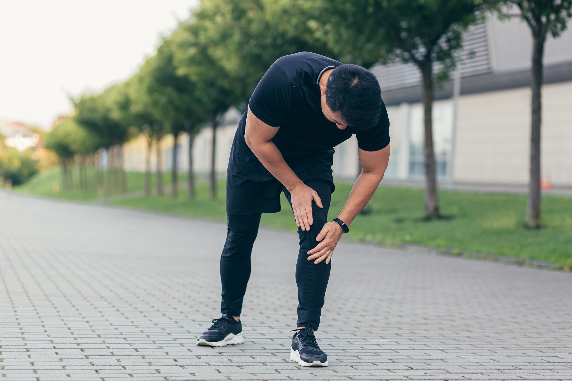 Man in black attire bends over, touching knee, possibly injured, on a paved path with trees in the background.