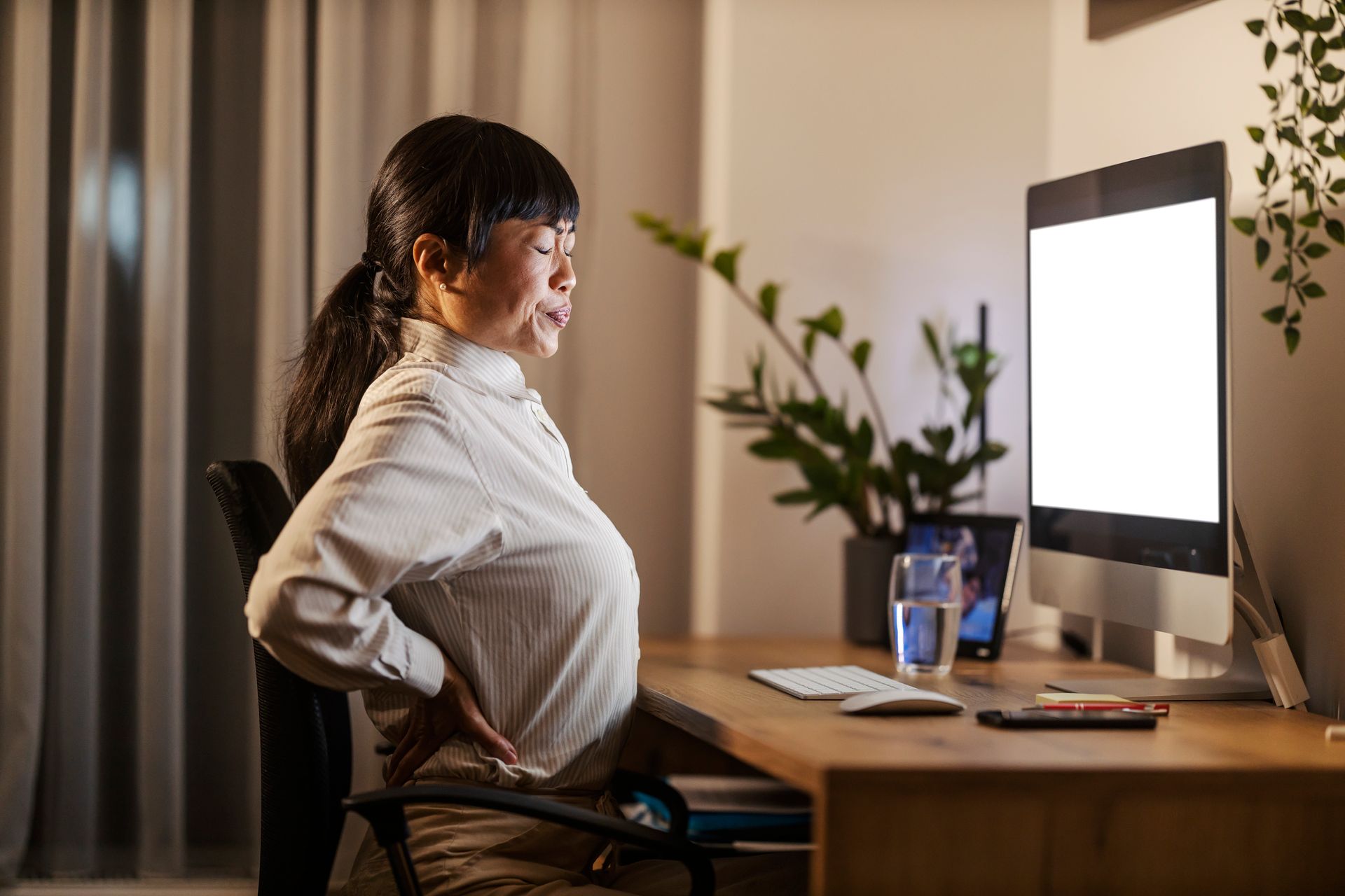 Woman at desk, holding her back in pain, looking at a computer.