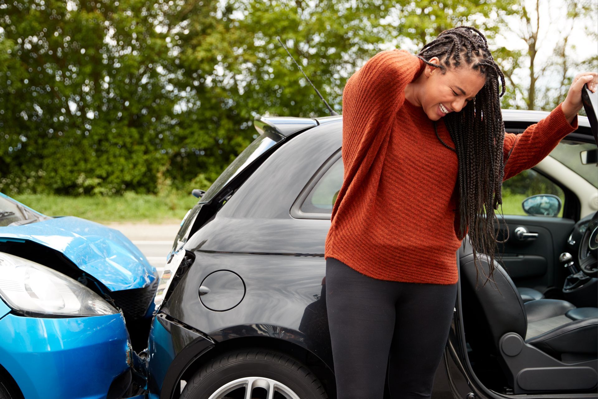 Woman exits a black car after a collision with a blue car; she holds her neck, appears in pain.