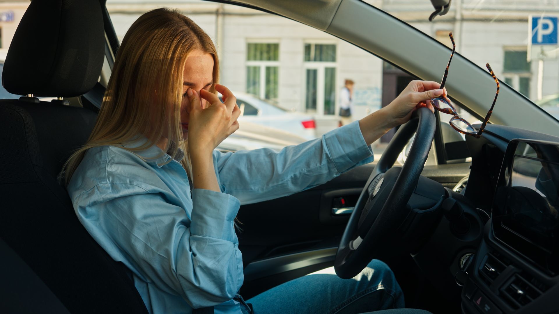 Woman in car, eyes closed, holding eyeglasses, appearing fatigued, daytime.
