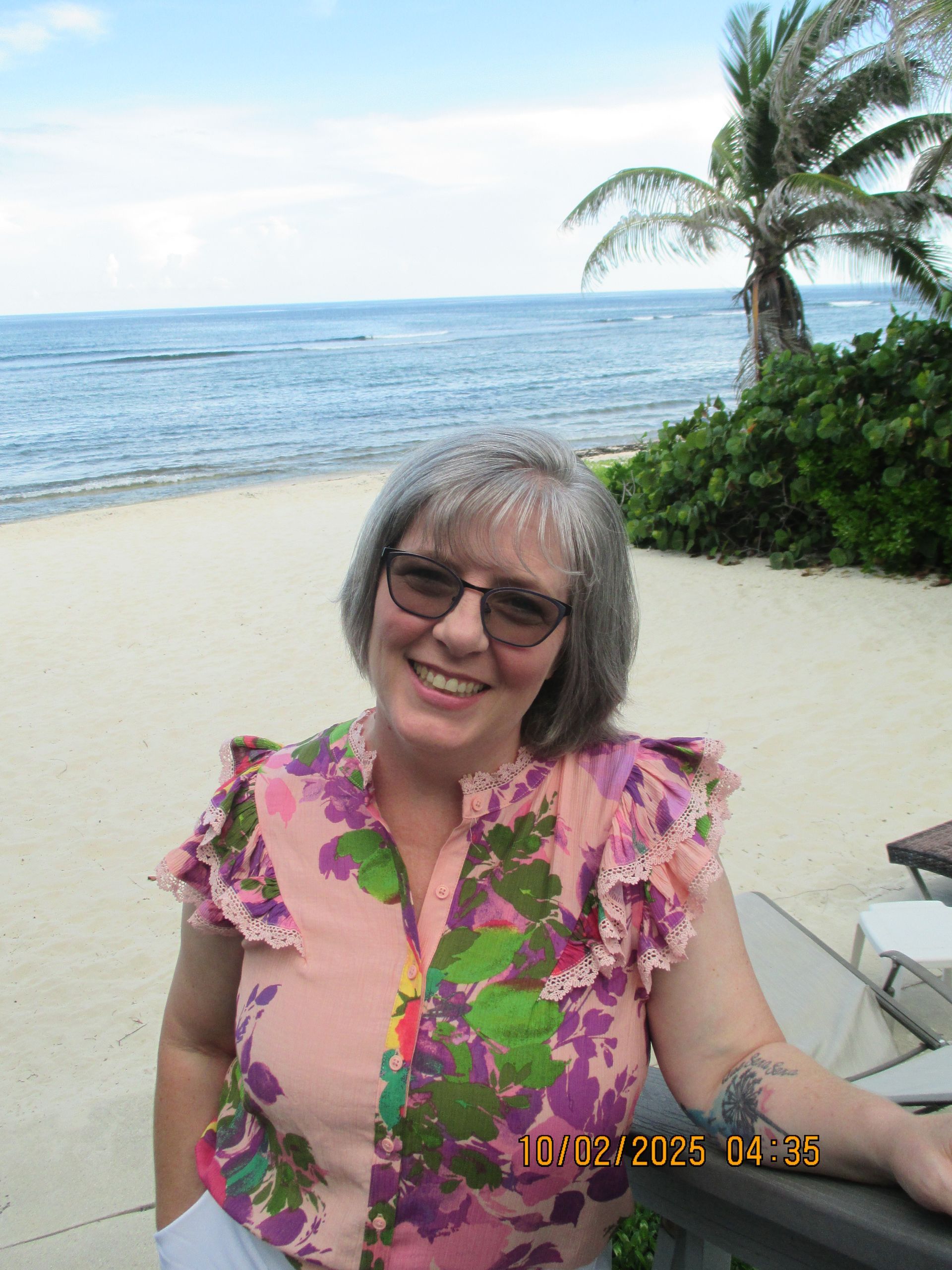 Woman smiles at the camera on a beach. She wears pink floral top, glasses; ocean and palm tree background.