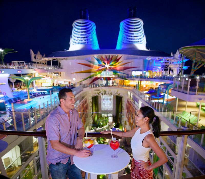Couple on cruise ship balcony with cocktails, ship lit up at night.